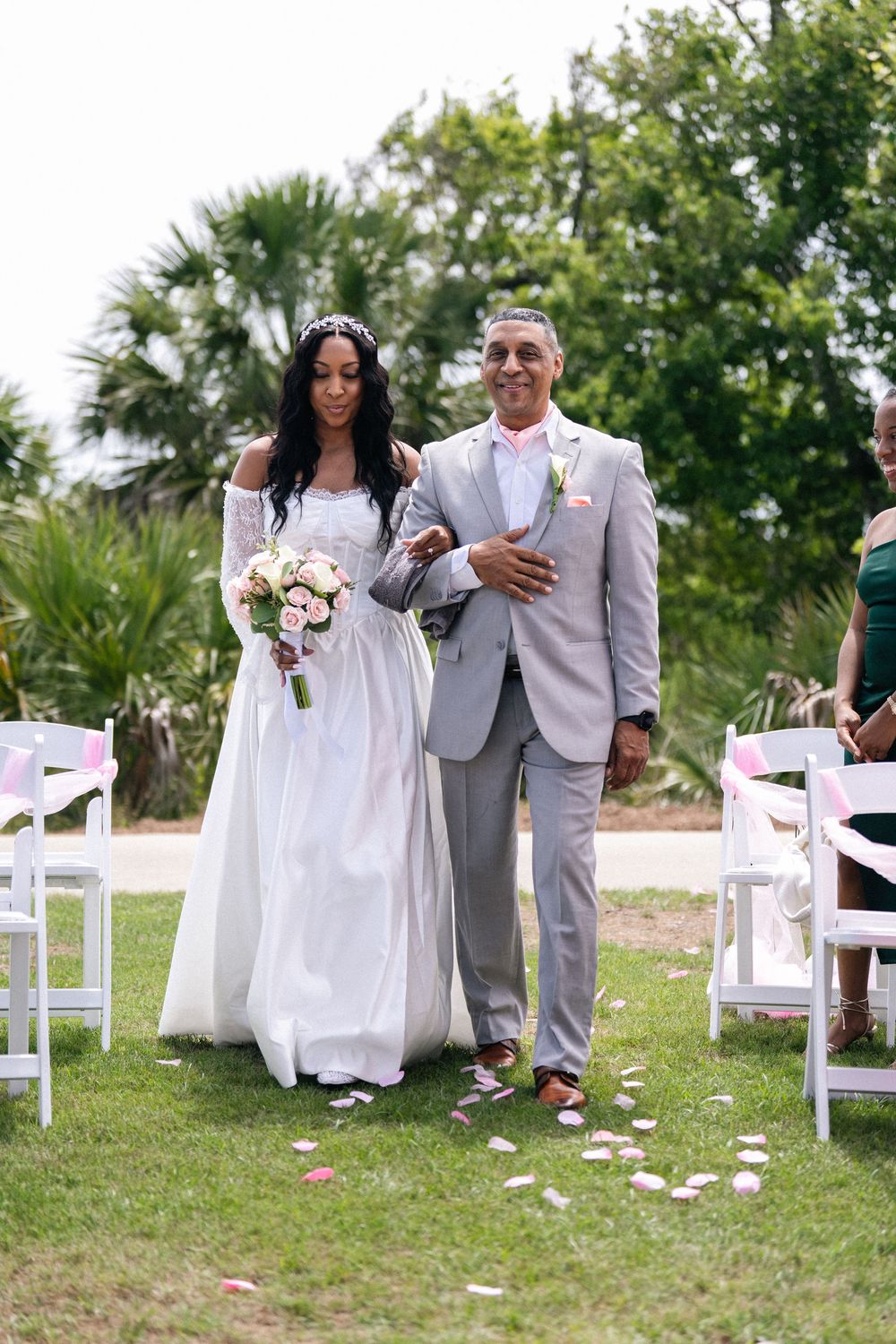 Bride in white gown holding pink bouquet walks down outdoor aisle with man in gray suit at garden wedding ceremony.