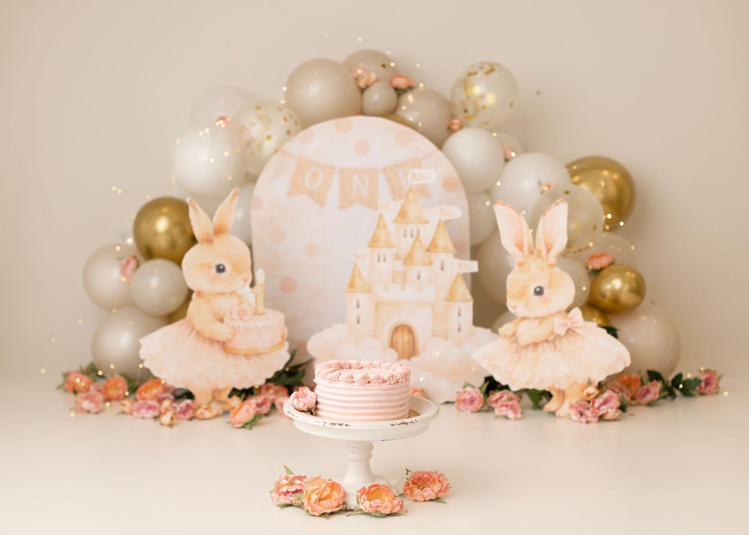 First birthday cake smash setup with bunny decor, balloon arch, pink florals, and a castle cake on white stand.