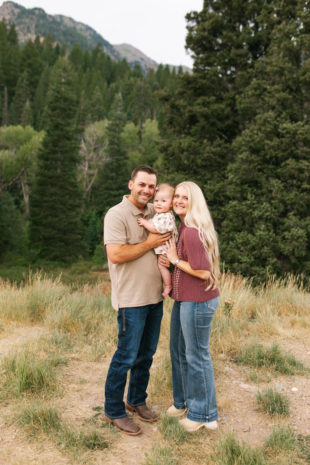 A smiling couple holds their baby outdoors near a mountain forest with tall evergreens and grassy meadow.