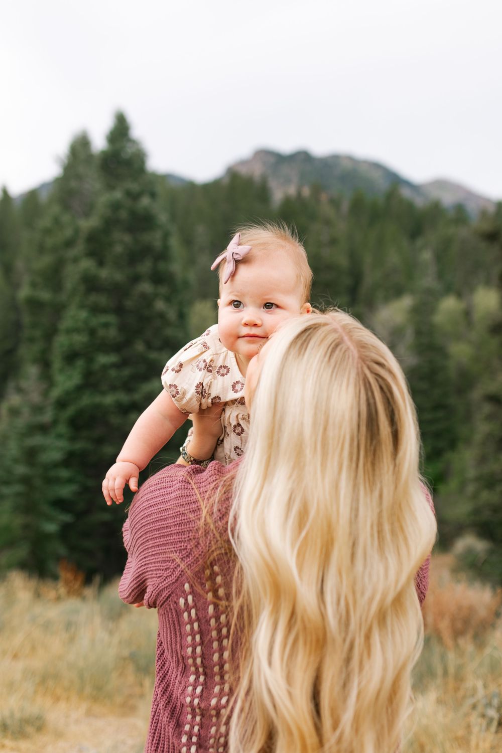Blonde mother lifting smiling baby girl with pink bow outdoors near pine trees and mountains.