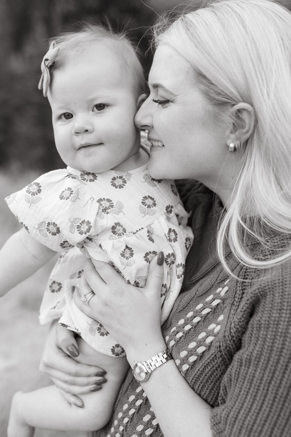 Black and white photo of a smiling blonde mother holding her baby girl in a floral dress outdoors.