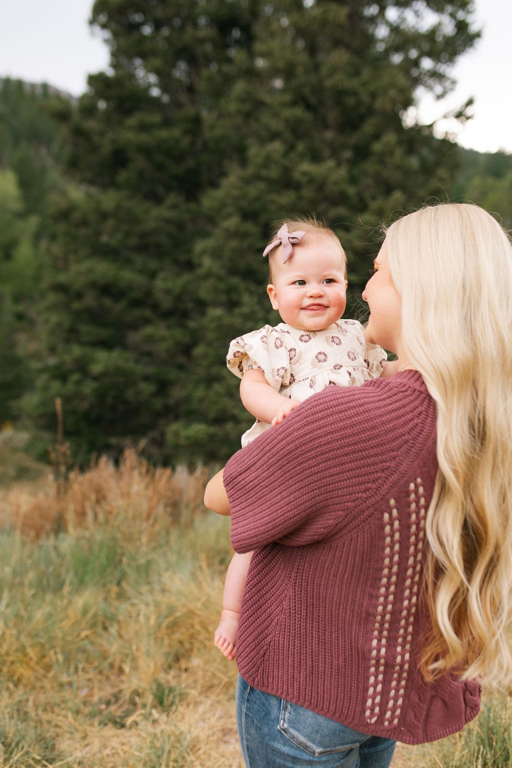 Blonde woman in burgundy sweater holding smiling baby girl in floral dress outdoors near evergreen trees.
