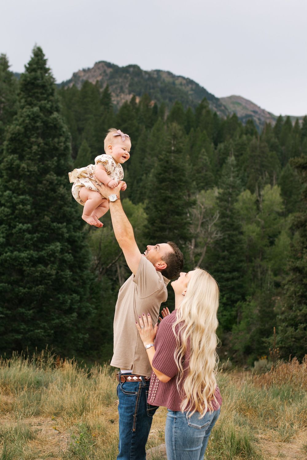 Father lifting baby girl overhead while mother watches, with pine trees and mountains behind.