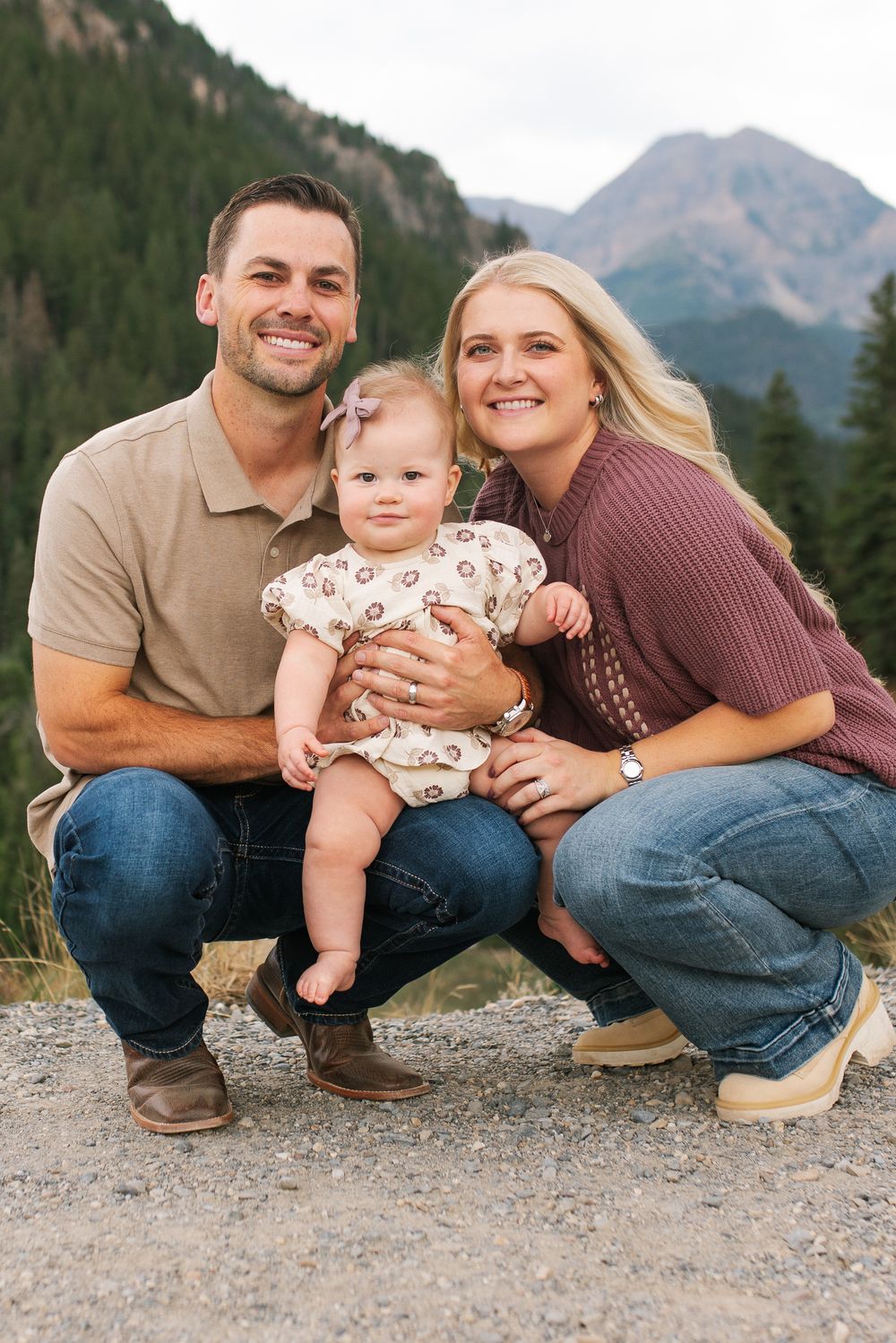 Smiling couple crouches with their baby girl in front of a scenic mountain landscape.