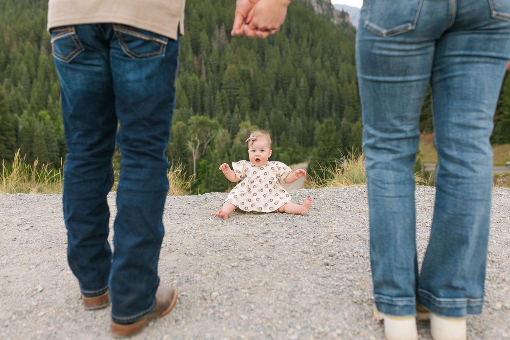 Baby sitting on gravel path between parents holding hands with mountain forest backdrop.