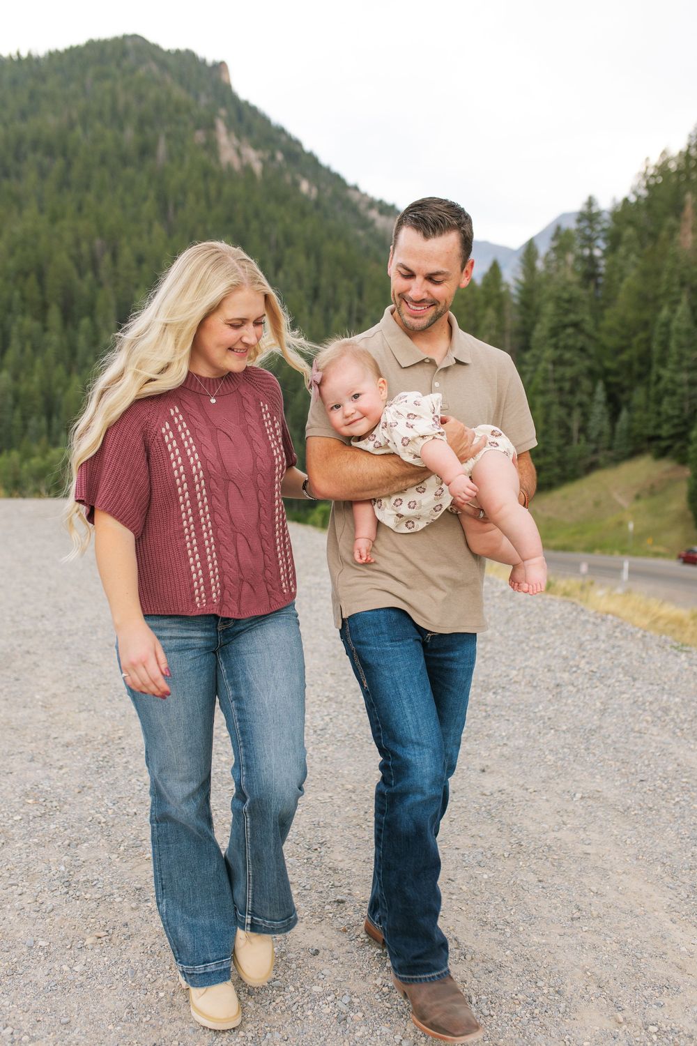 A happy young family with a smiling man holding a baby and a blonde woman walking on a gravel path with mountains behind.