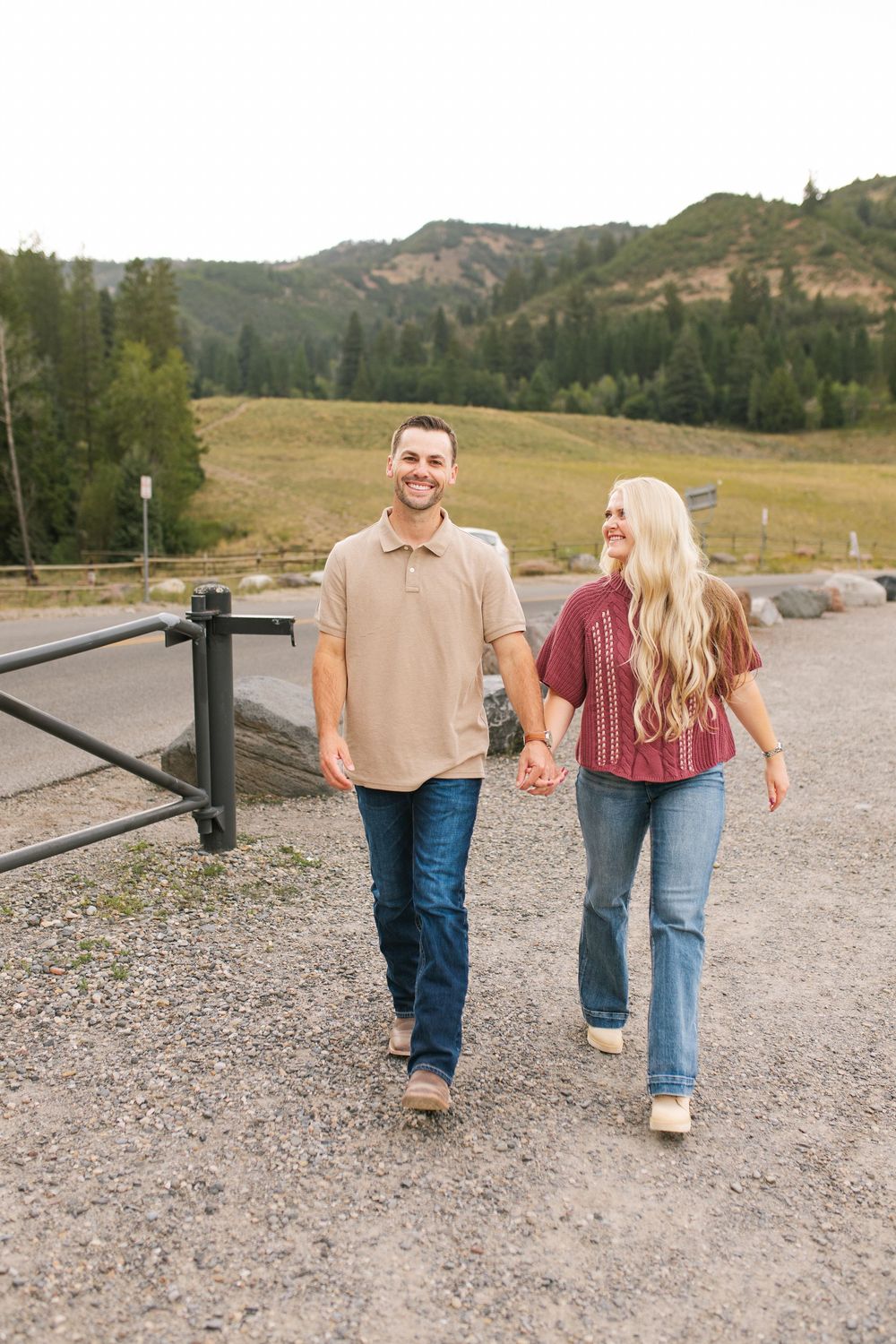Happy couple holding hands walking on gravel path with green mountain landscape in background.
