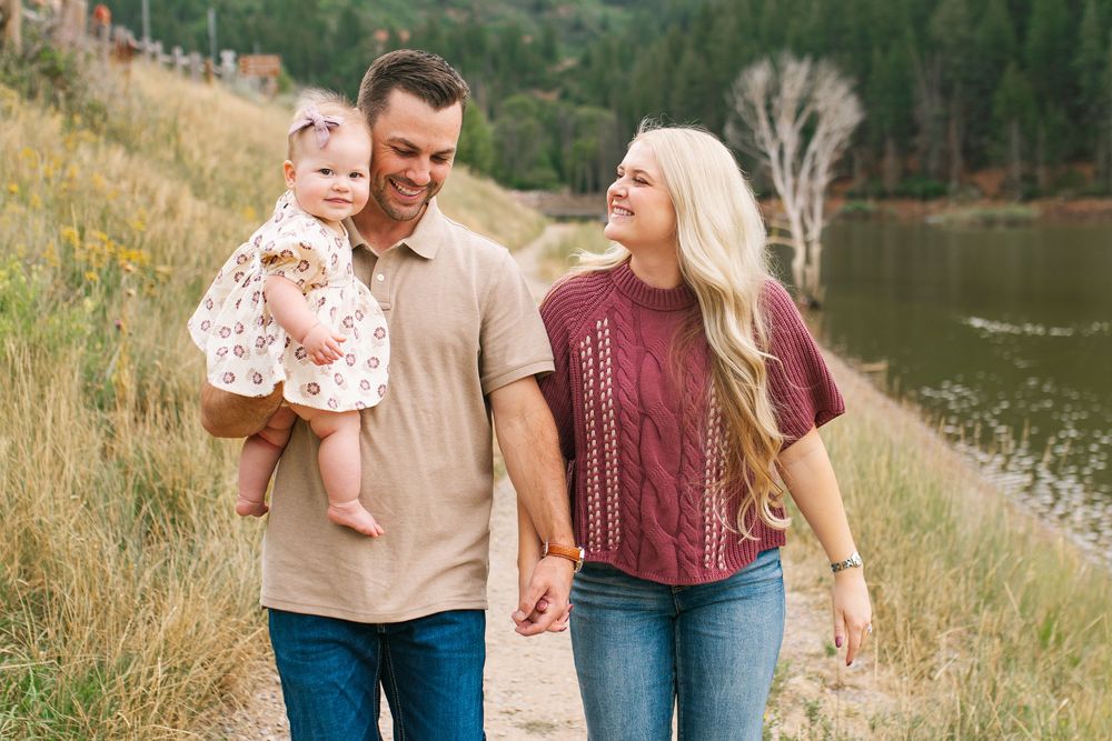 Happy family of three walking outdoors near a mountain lake, parents smiling at baby girl in floral outfit.