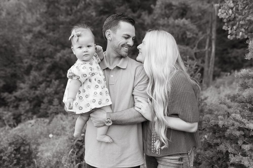 Black and white family portrait of a man holding a baby girl while a blonde woman leans in, outdoors.