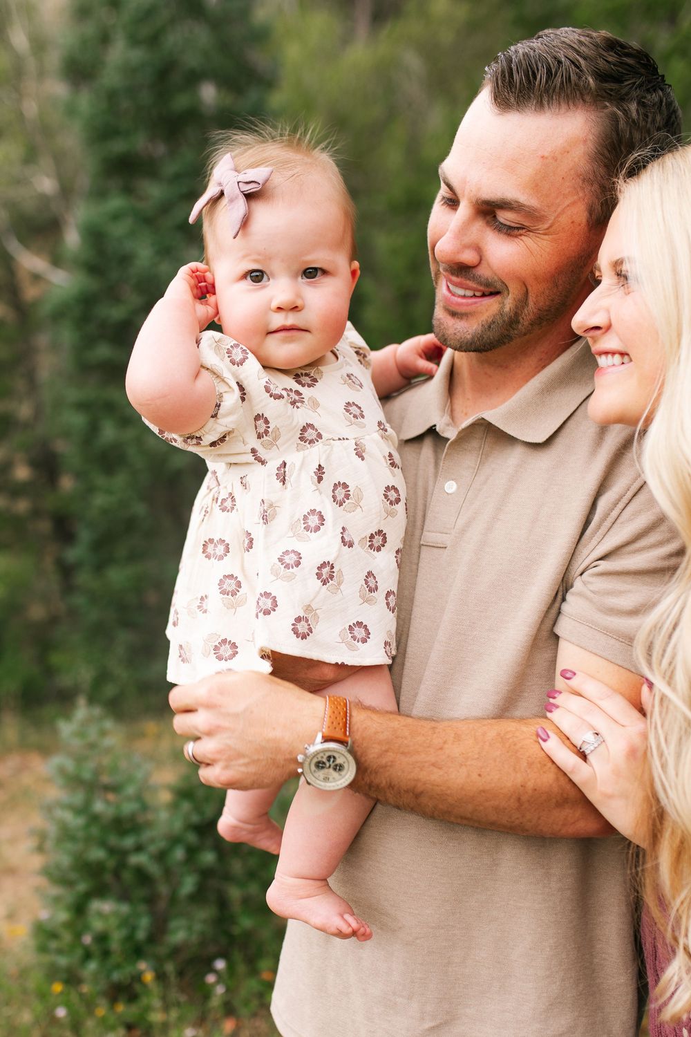 Smiling parents holding adorable baby girl with bow outdoors in green forest setting.