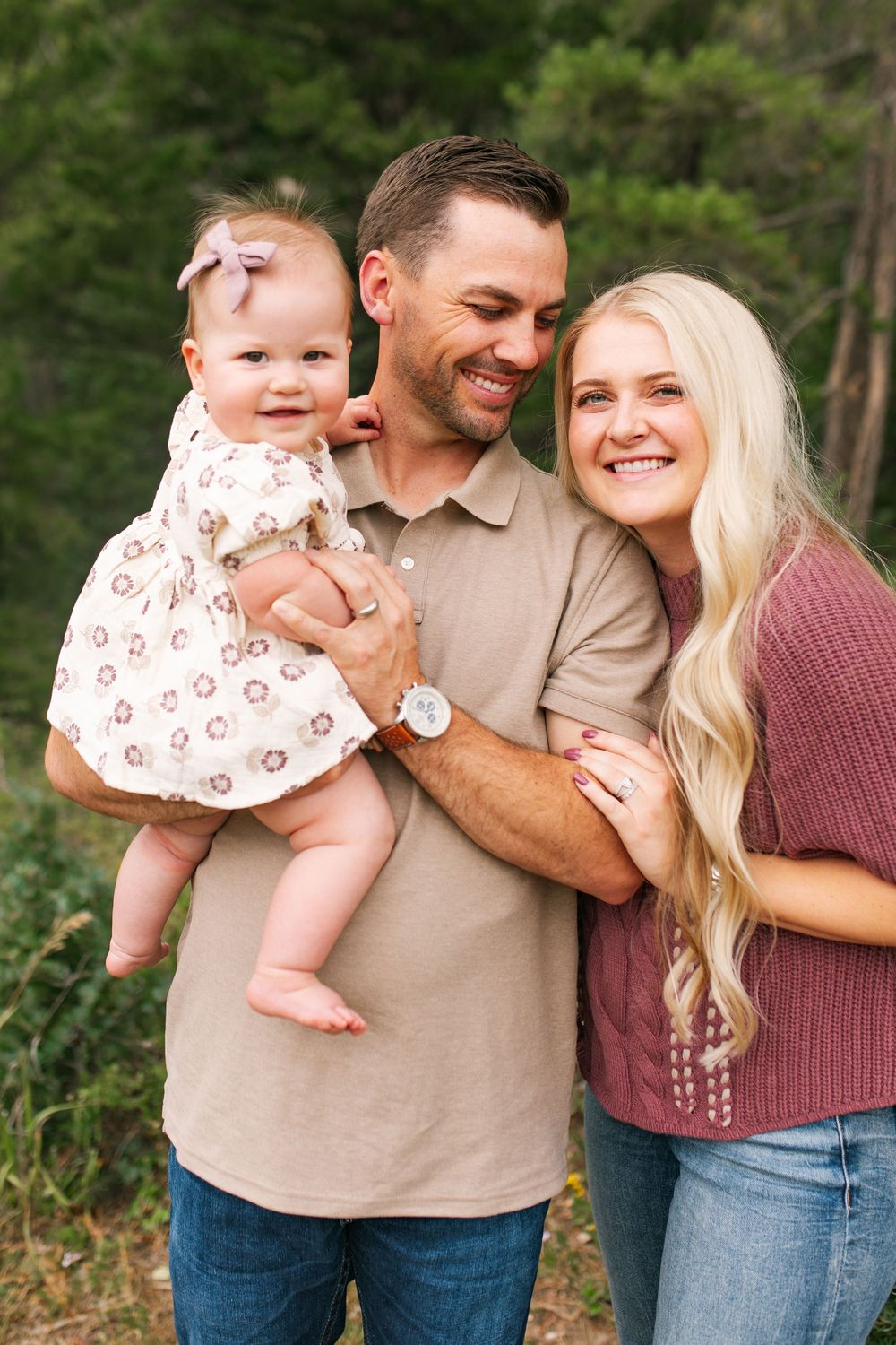 Smiling family with man holding baby girl in floral dress, woman leaning close, outdoors with green trees.