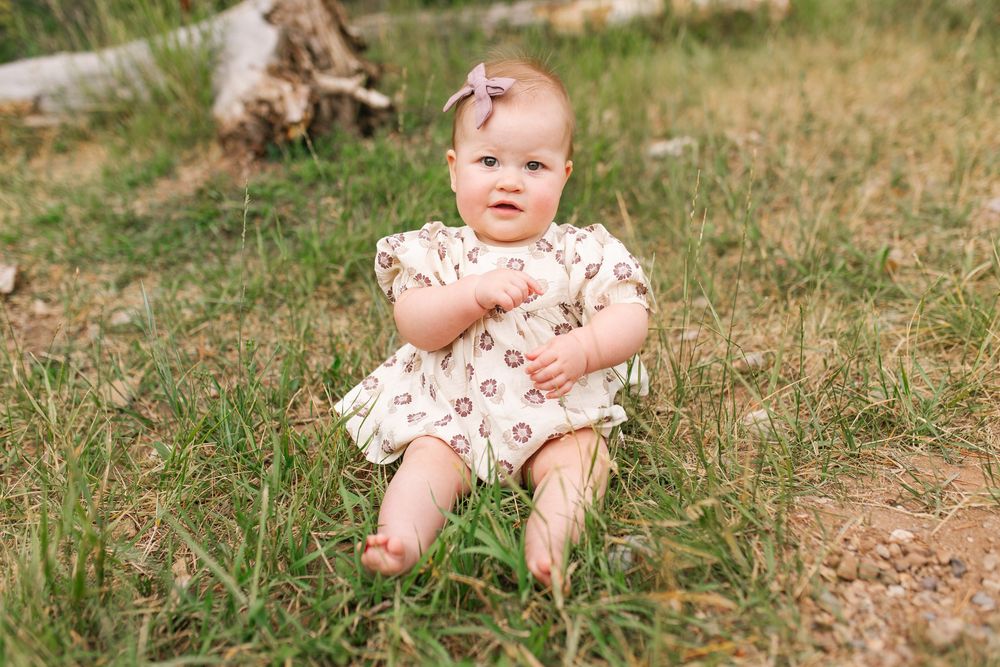 Baby girl in floral romper with bow headband sitting on green grass outdoors.