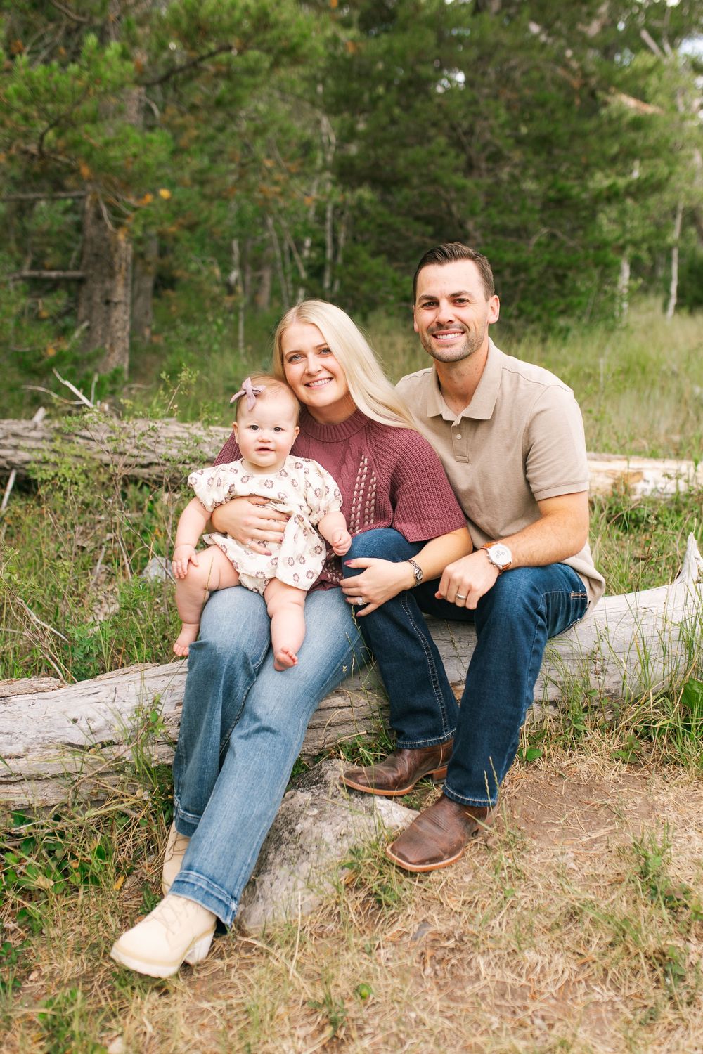 A smiling young family with a baby girl sits on a log in a lush green forest setting for a portrait.