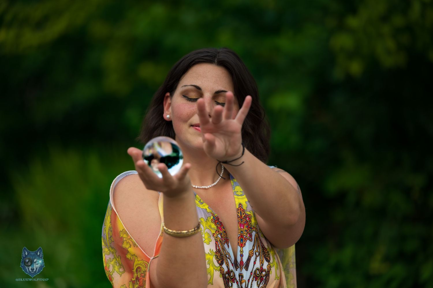 Woman in floral top balances crystal ball on palm with eyes closed in green garden setting. Chi.Shooters meet gabbsbosh