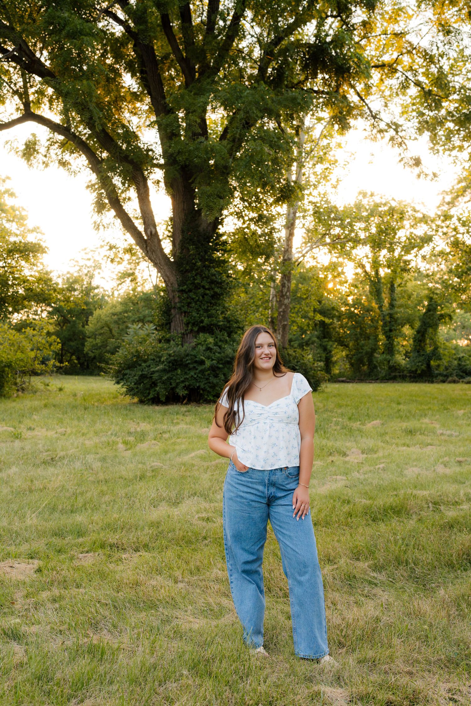 Young woman in jeans and floral top smiling outdoors at golden hour with large tree and green grass.
