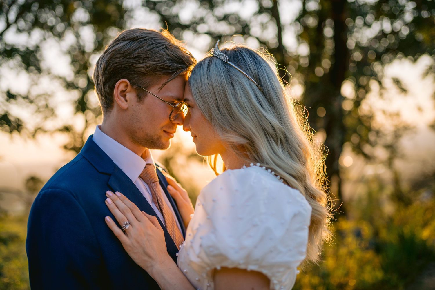 A couple shares a romantic moment at golden hour, the bride in white and groom in navy suit, with warm sunset light glowing behind them.