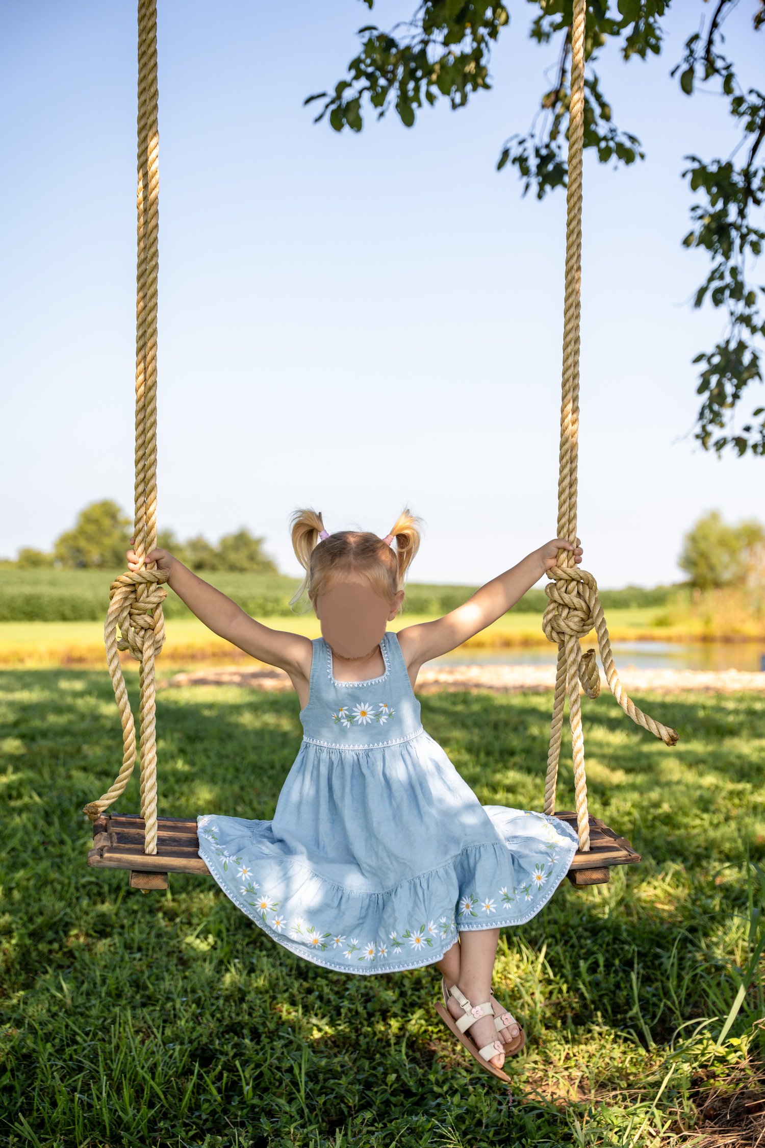A young girl in a blue floral dress sits on a rope swing outdoors, arms raised, enjoying a sunny day in a green park.