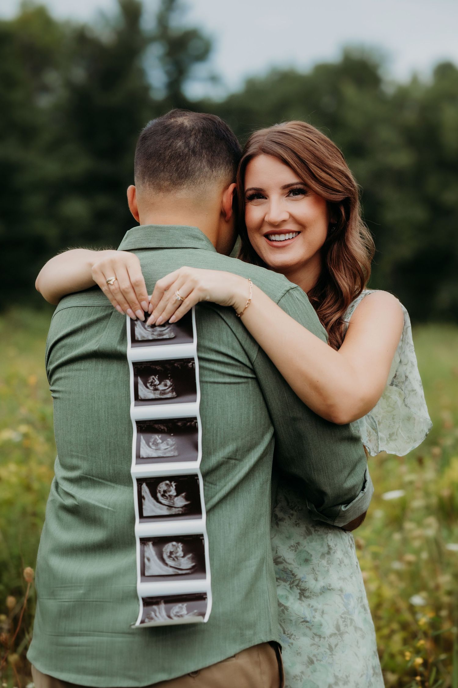 Smiling woman in floral dress hugs man outdoors holding ultrasound photos, announcing pregnancy in a green field.