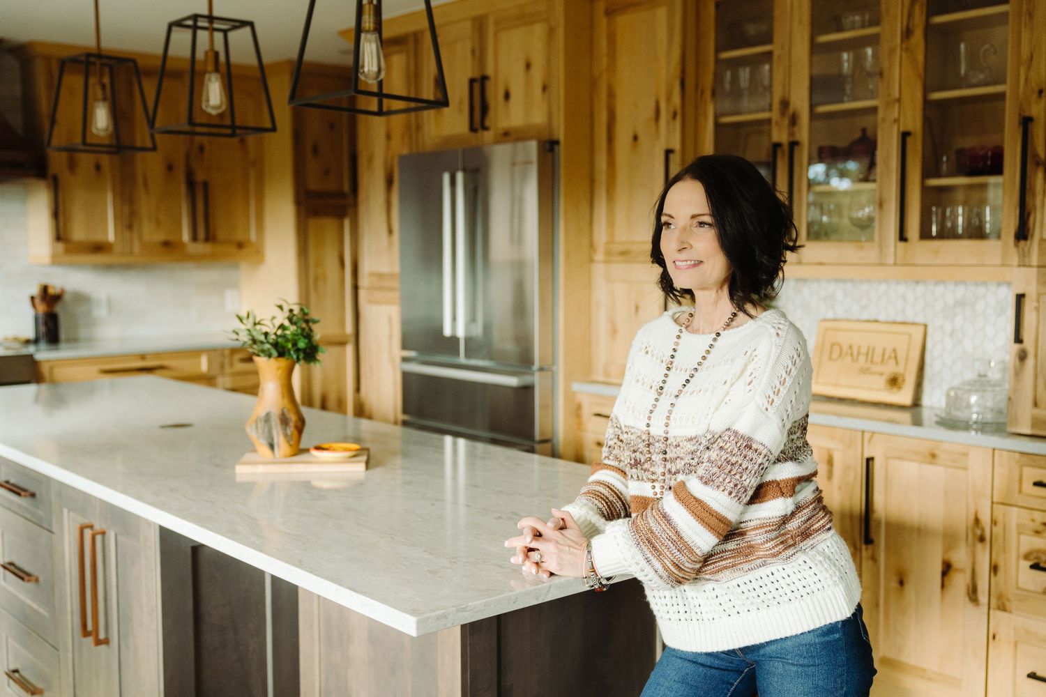 Woman in cozy sweater leaning on kitchen island in warm wood cabin kitchen with stainless steel appliances.