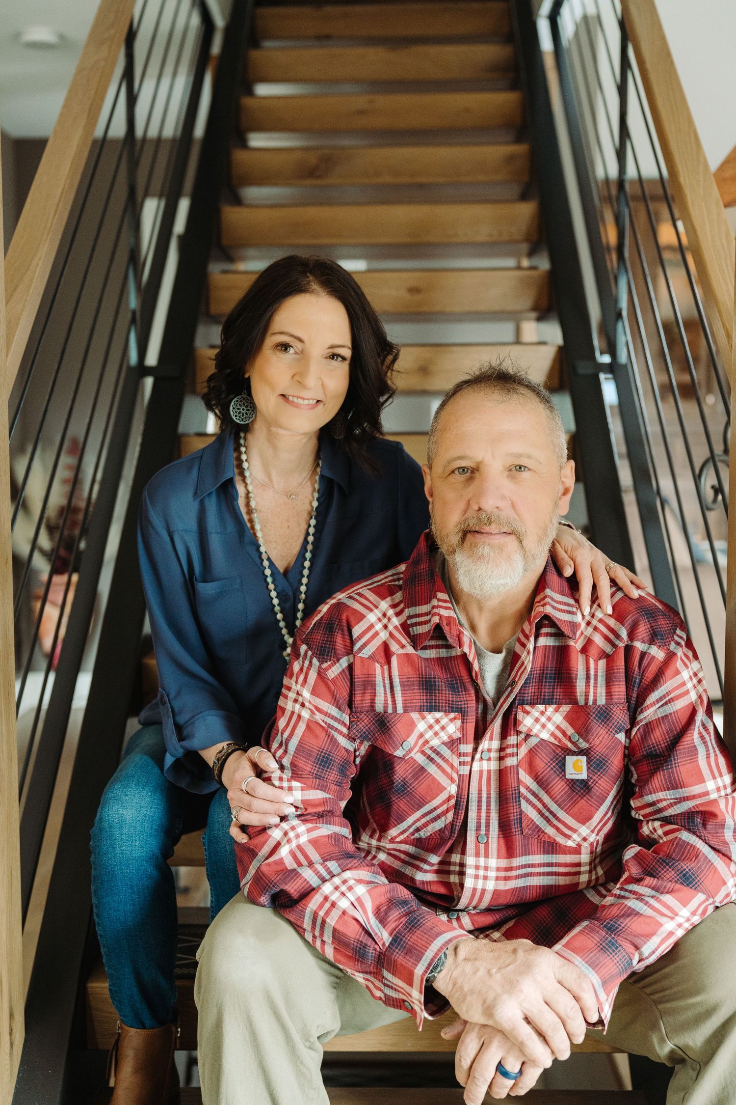 A middle-aged couple sitting together on wooden stairs, man in plaid Carhartt shirt, woman in navy blouse with pearl necklace.