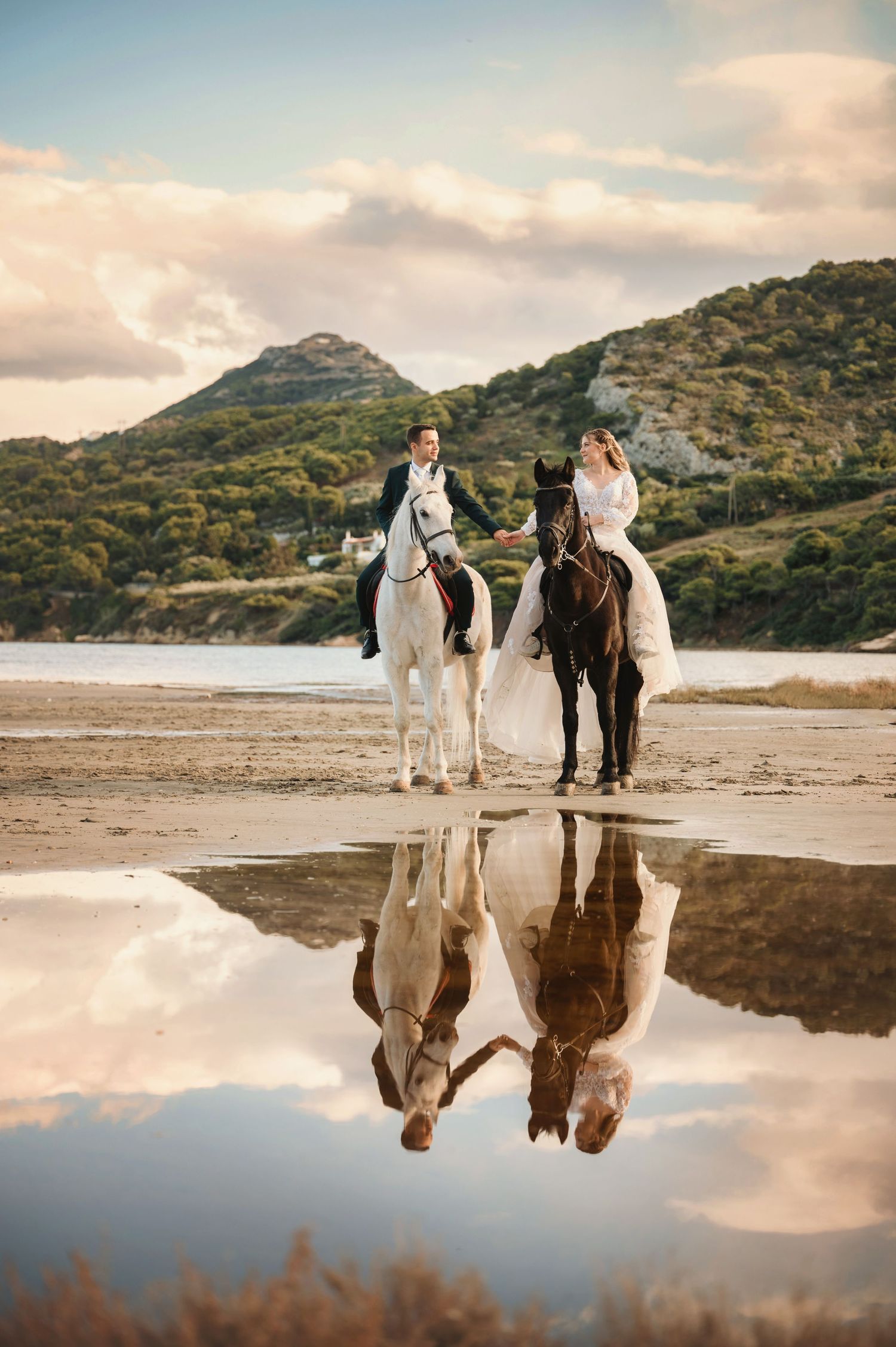 Couple in wedding attire riding horses on a beach with mountain backdrop and reflections in water.