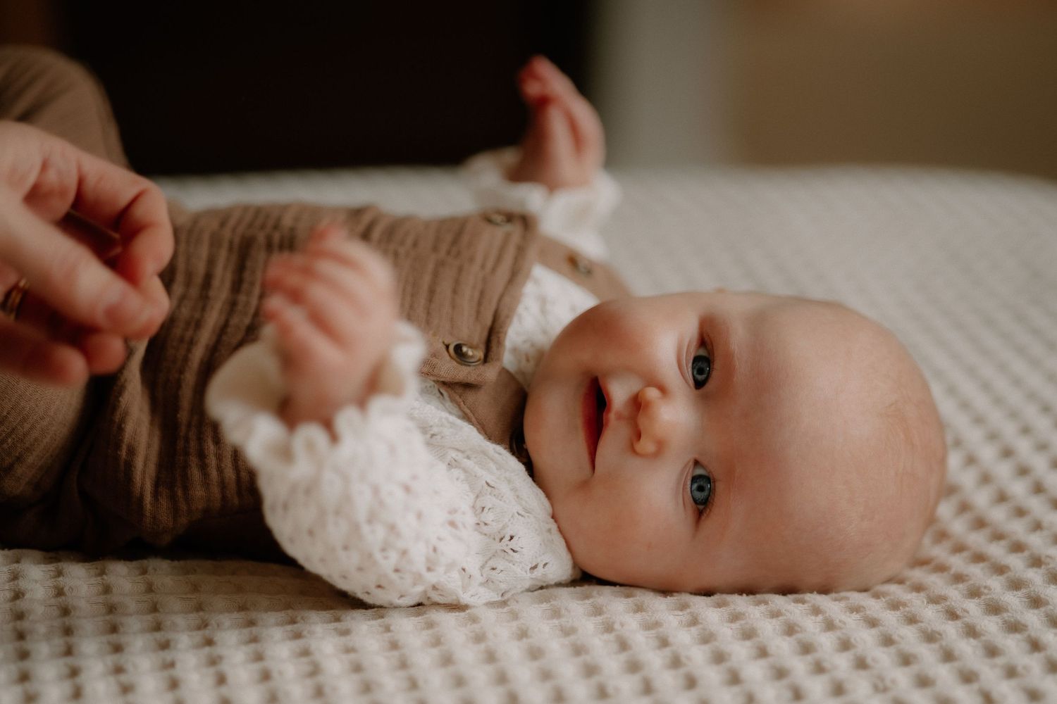 A newborn baby in a brown knit outfit lies on a white textured blanket, gazing curiously with bright blue eyes.