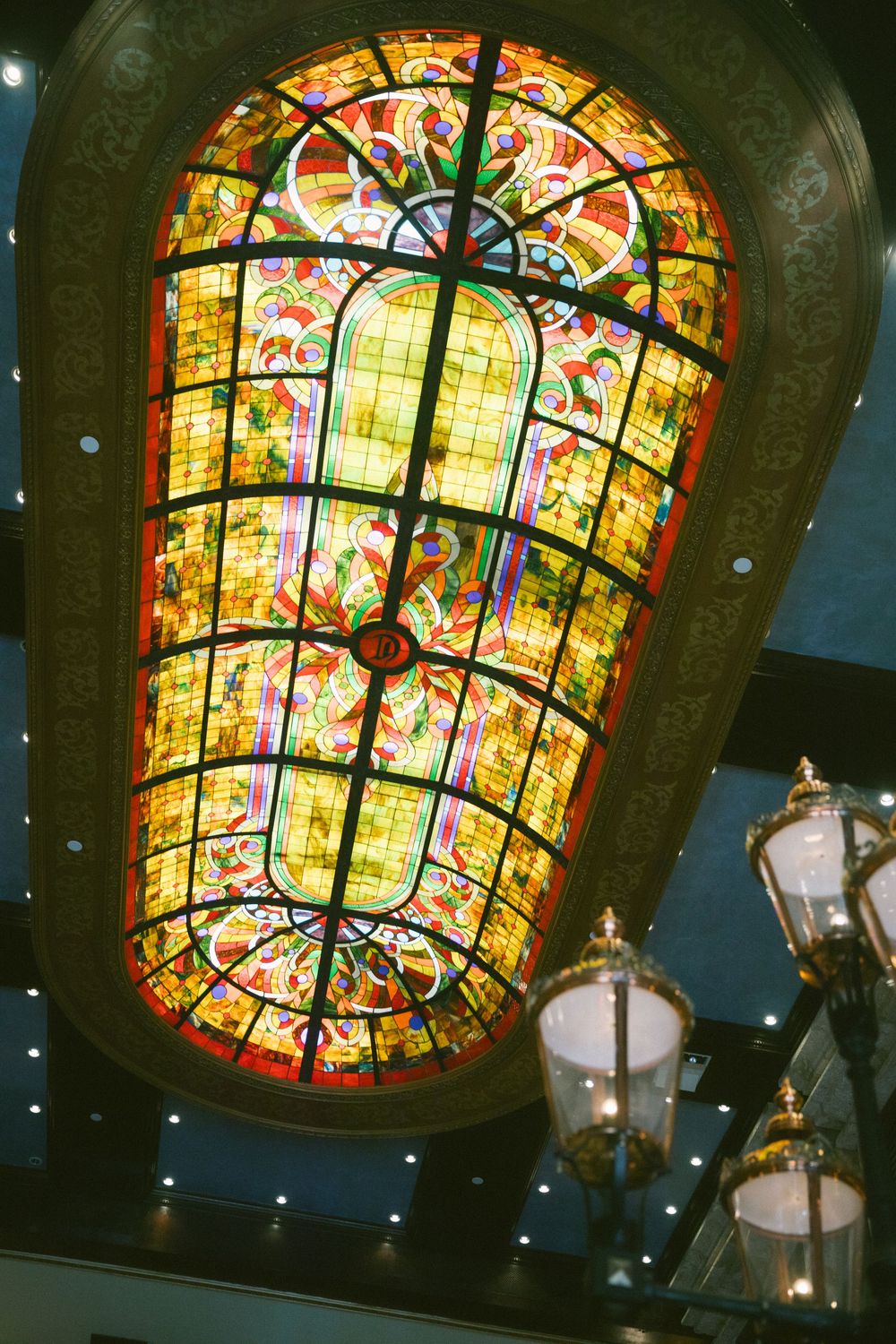 Ornate stained glass skylight ceiling with colorful floral patterns and elegant chandelier in a grand interior.