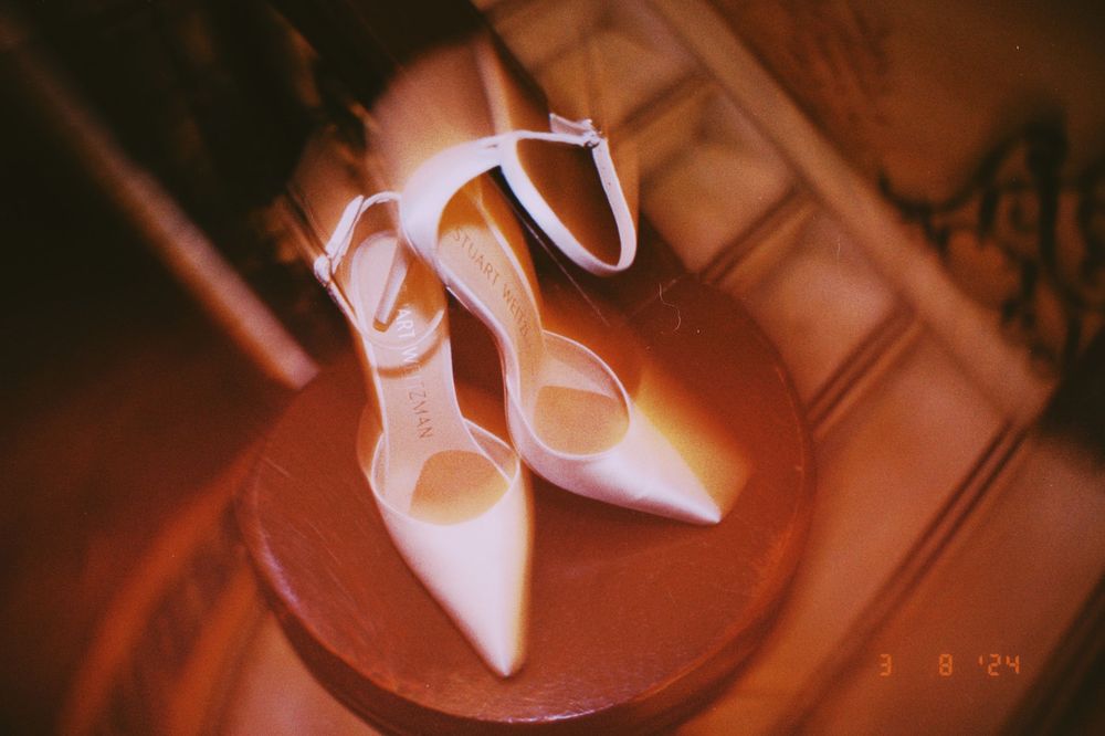 Elegant white pointed-toe heels with ankle straps displayed on a round wooden stand in warm lighting.