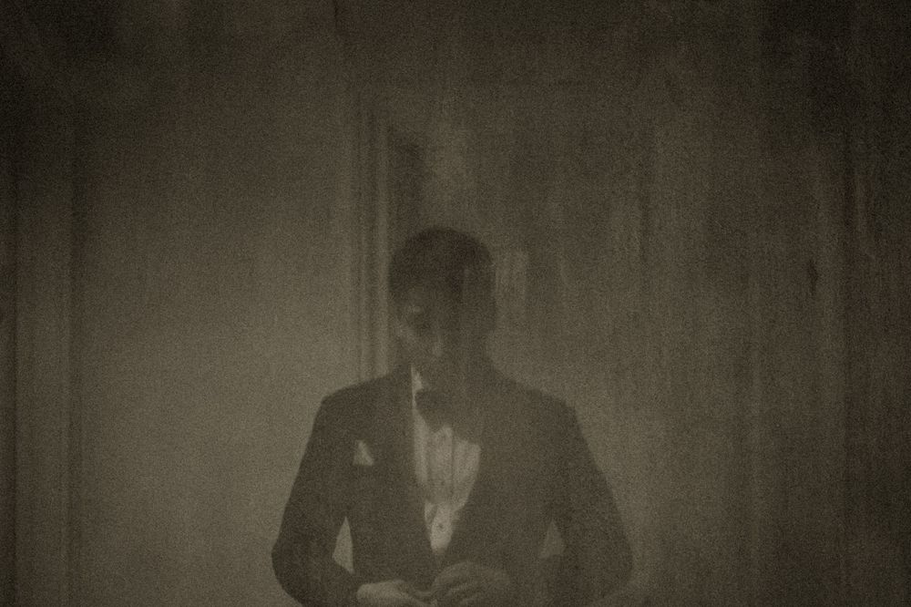A mysterious man in a suit stands in dim, moody lighting in a dark, vintage-style atmospheric photograph.