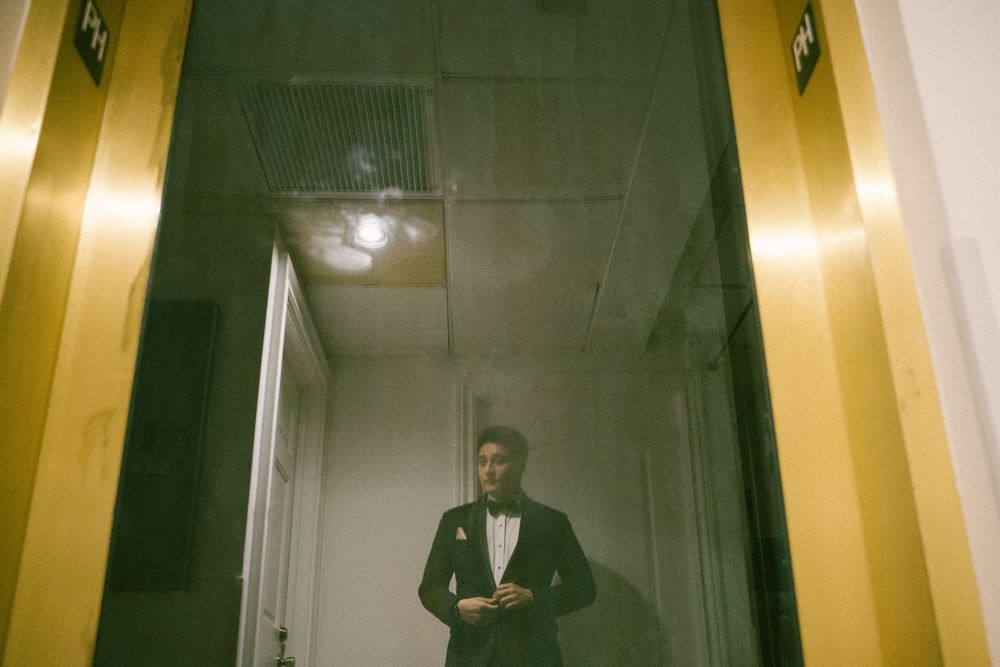 Elegant man in black tuxedo standing in golden elevator doorway at formal event.