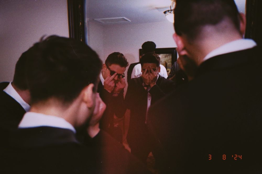 Group of people in formal black attire gathered closely together in a dimly lit room, date stamped 3.8.24.