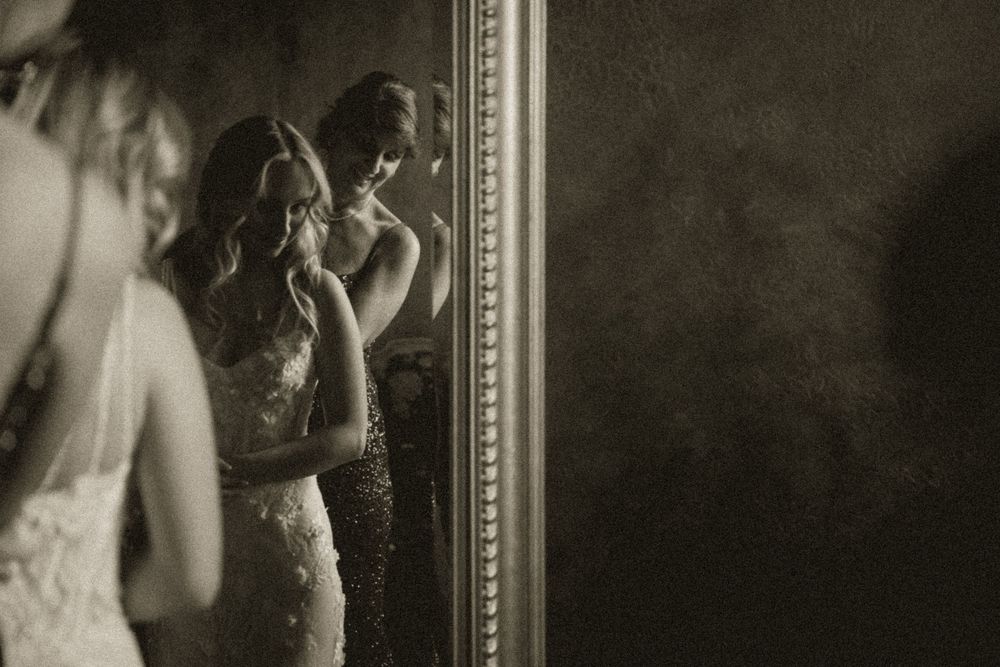 Bride in lace wedding gown gazes into ornate mirror in moody black and white portrait.