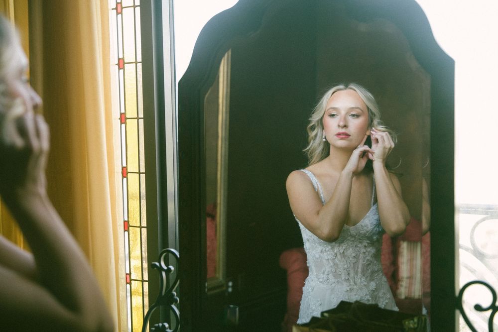 Blonde bride putting on earrings while looking in an ornate mirror during wedding day preparations.