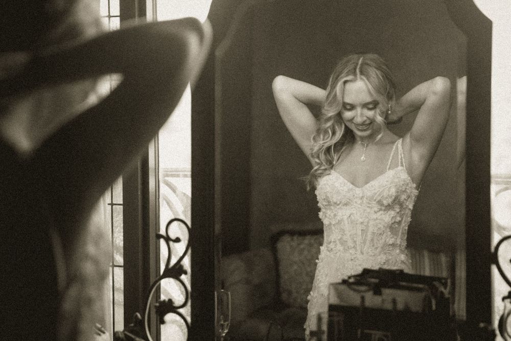 Bride in lace corset dress adjusts hair while gazing at her reflection in an ornate mirror, black and white photo.