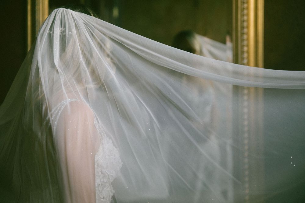 Bride's white wedding veil dramatically draped over her head, reflected in a gold-framed mirror.