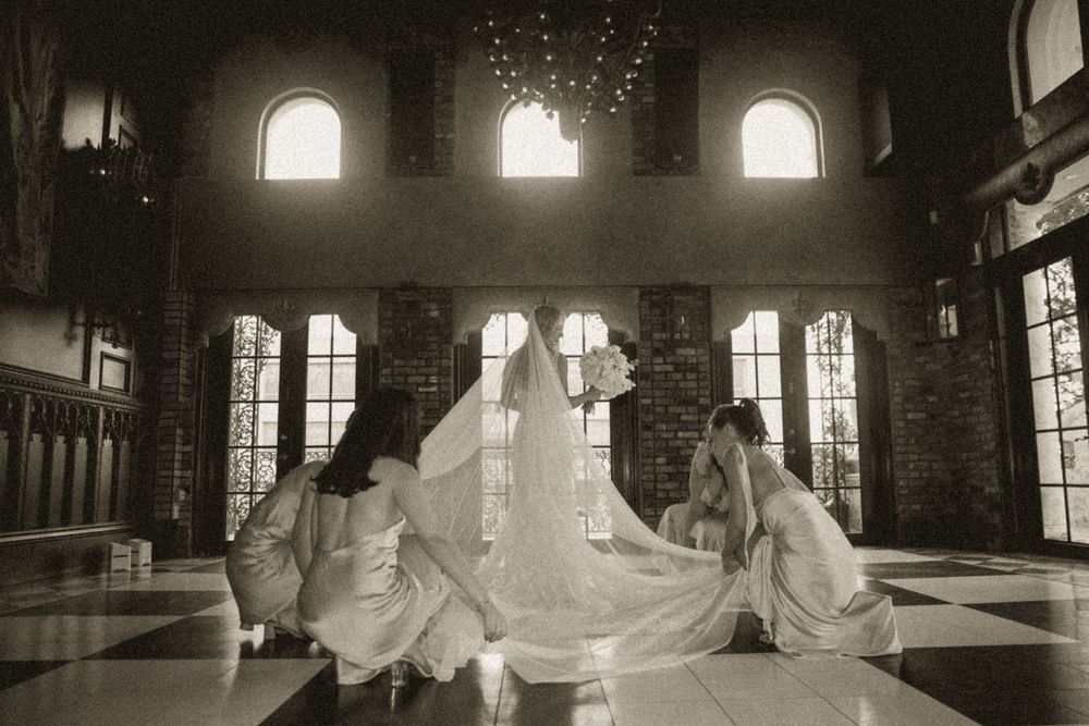 Black and white photo of a bride and bridesmaids arranging a wedding veil in an elegant hall.