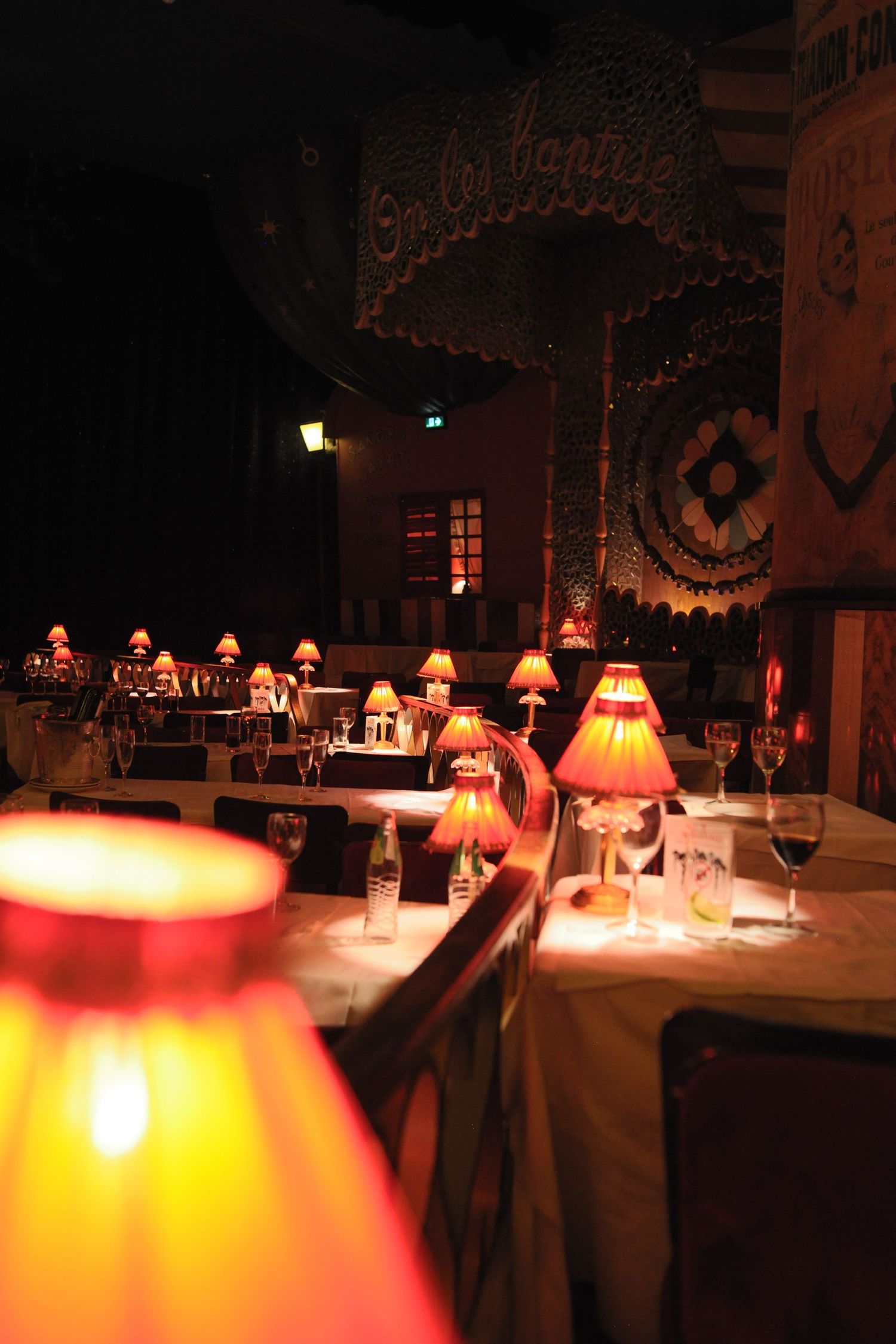 Dimly lit restaurant with warm glowing table lamps, white tablecloths, wine glasses, and decorative vintage signage in background.
