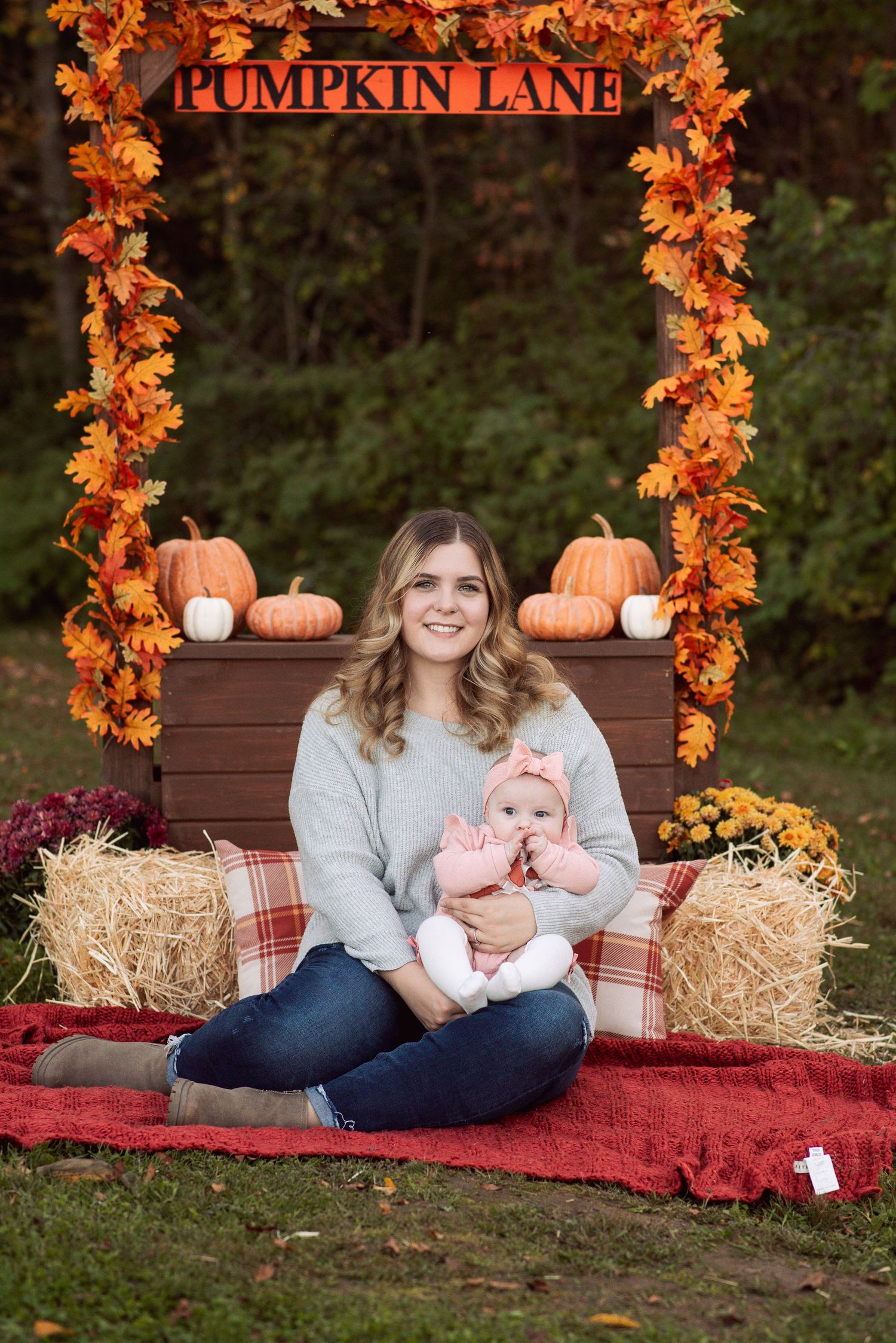 Woman sitting cross-legged on red blanket holding baby at fall Pumpkin Lane display with hay bales and pumpkins.