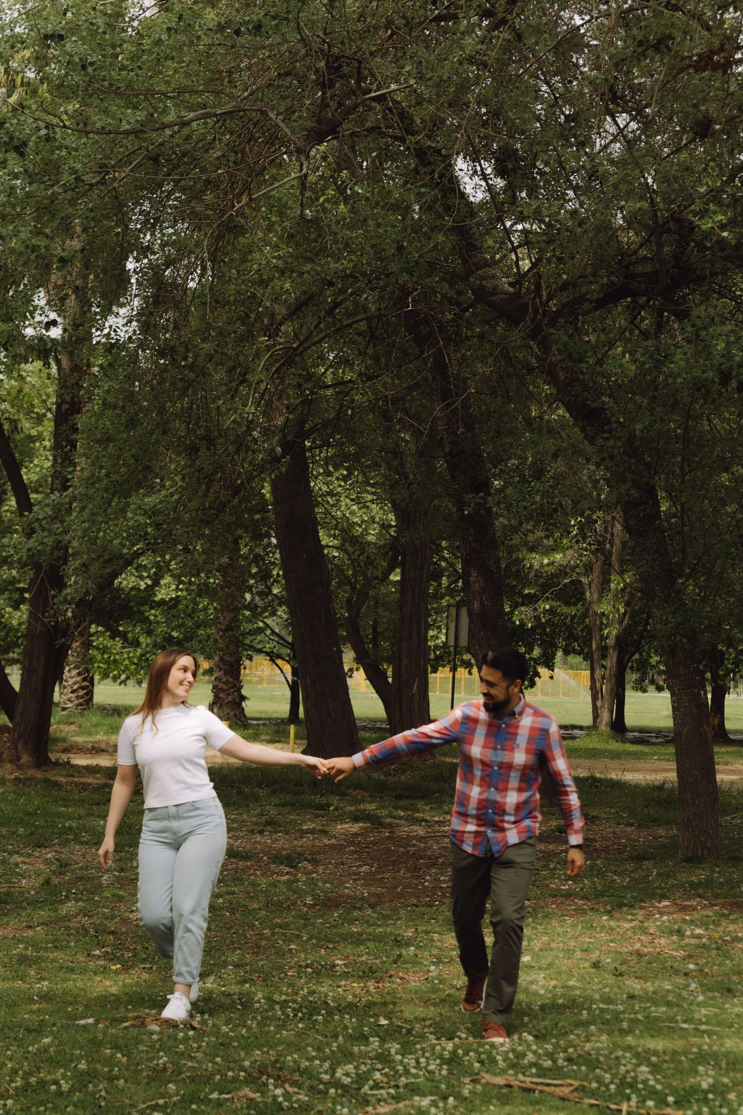 Happy couple holds hands walking through tree-lined green park on a cloudy day.