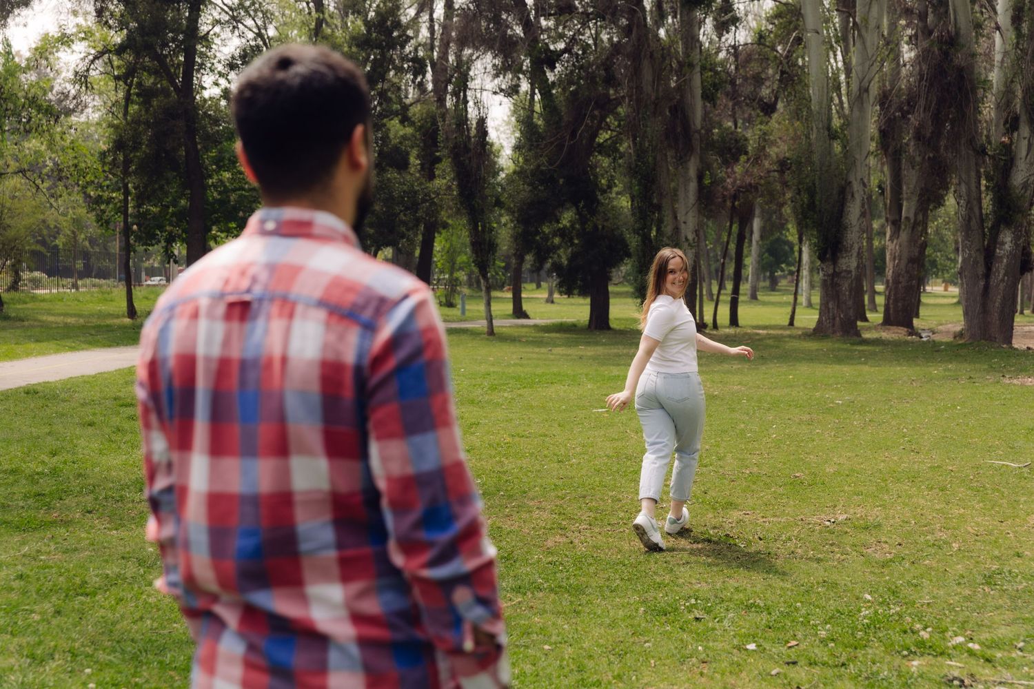 Young woman in white top and jeans walks away from man in plaid shirt in park.