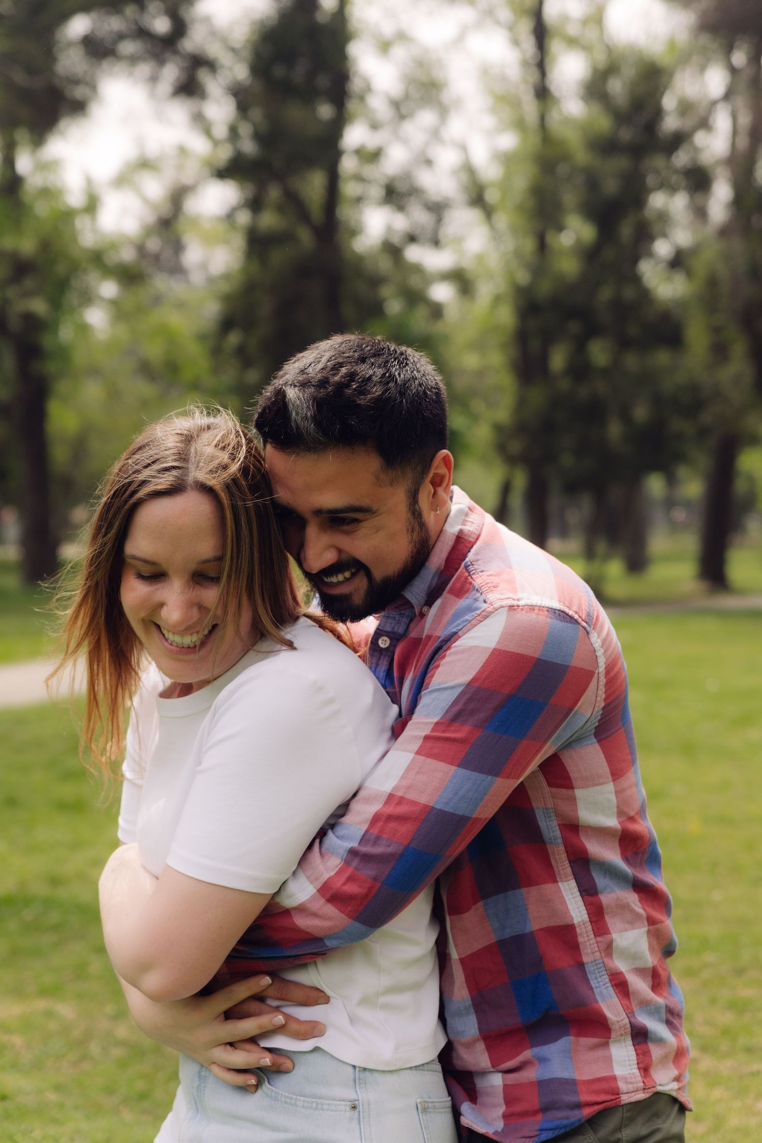 Happy couple embracing and laughing together in a green park, man in plaid shirt hugging woman from behind.