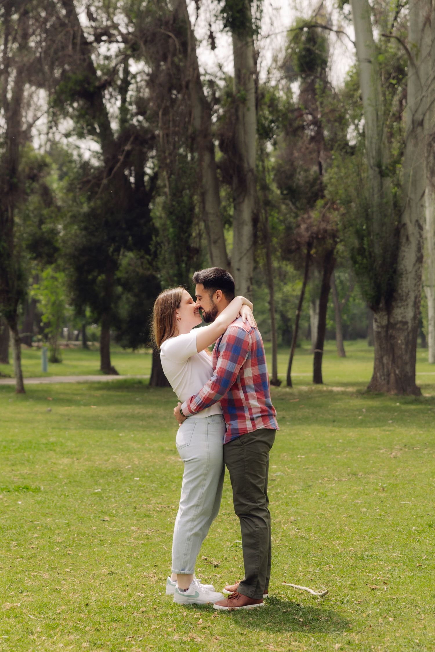 Romantic couple sharing a kiss while standing on green grass in a tree-lined park on a cloudy day.