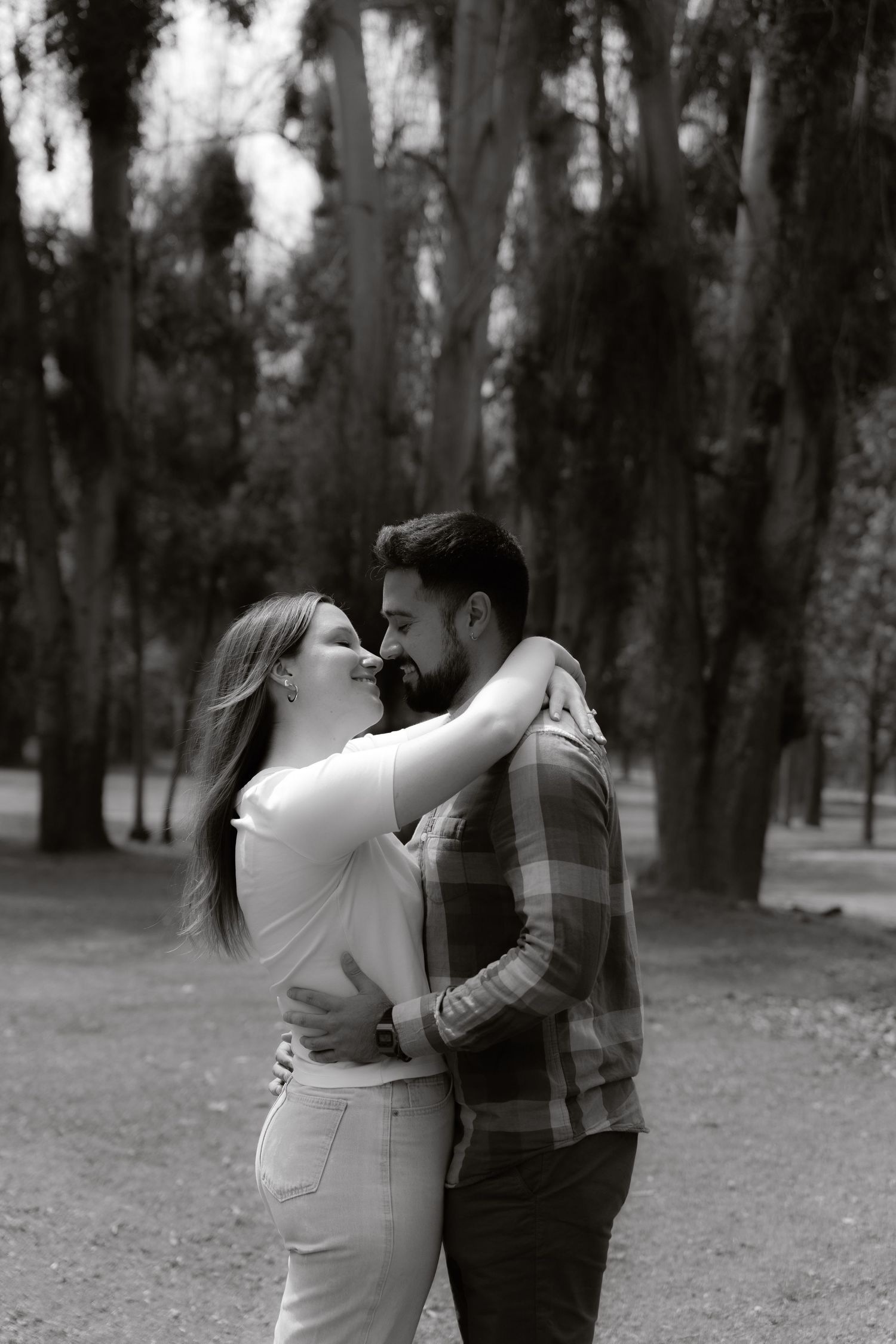 Black and white photo of a loving couple embracing and gazing at each other among tall trees in a park.