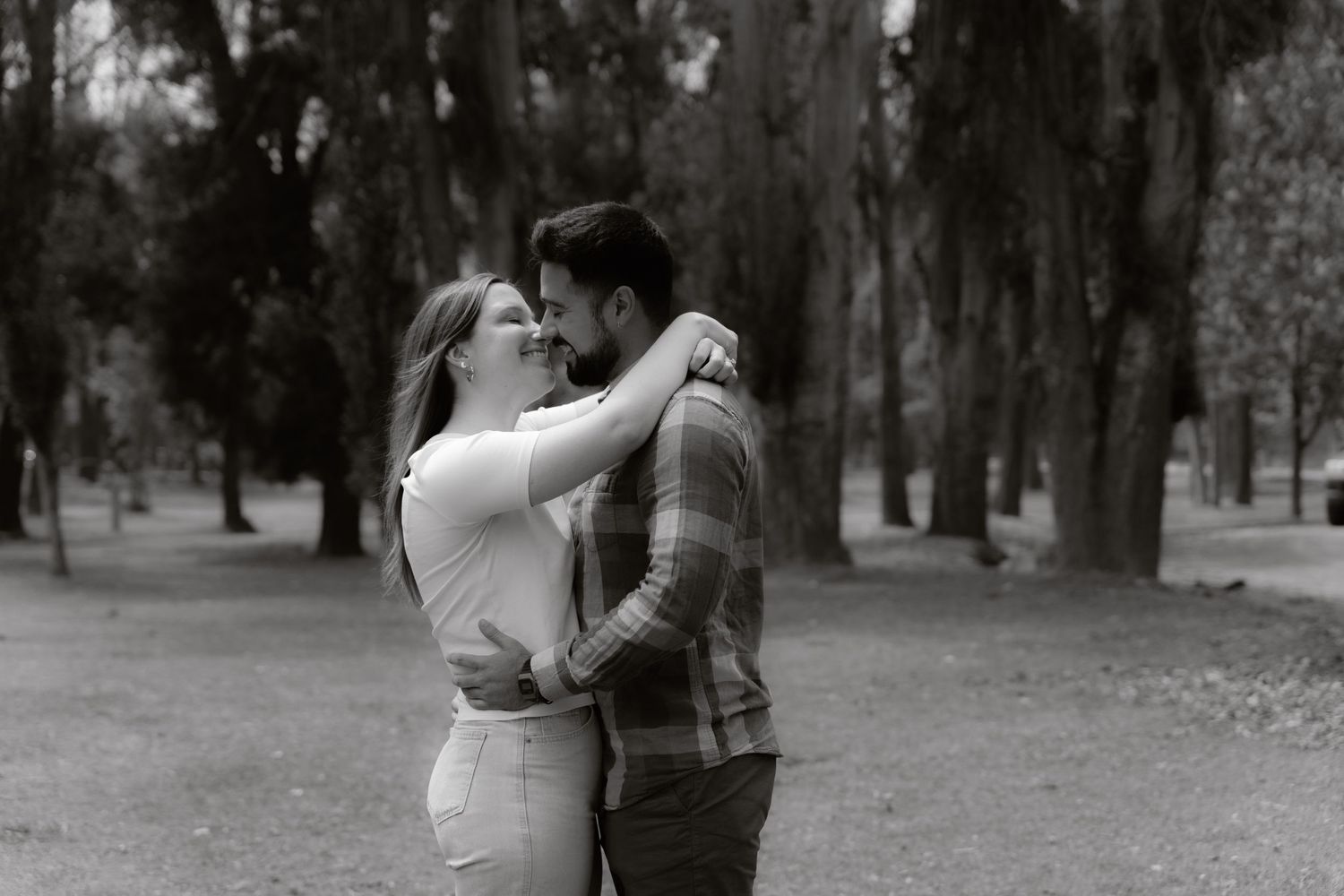 Black and white romantic photo of a couple nearly kissing while embracing outdoors in a wooded park setting.