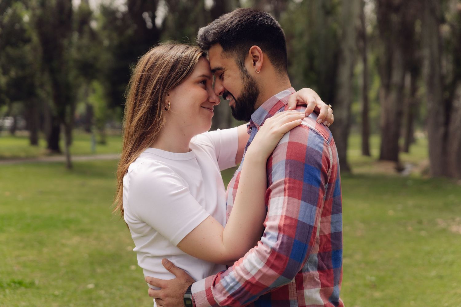 Affectionate couple touching foreheads and embracing in a lush green park, woman in white top and man in plaid shirt.