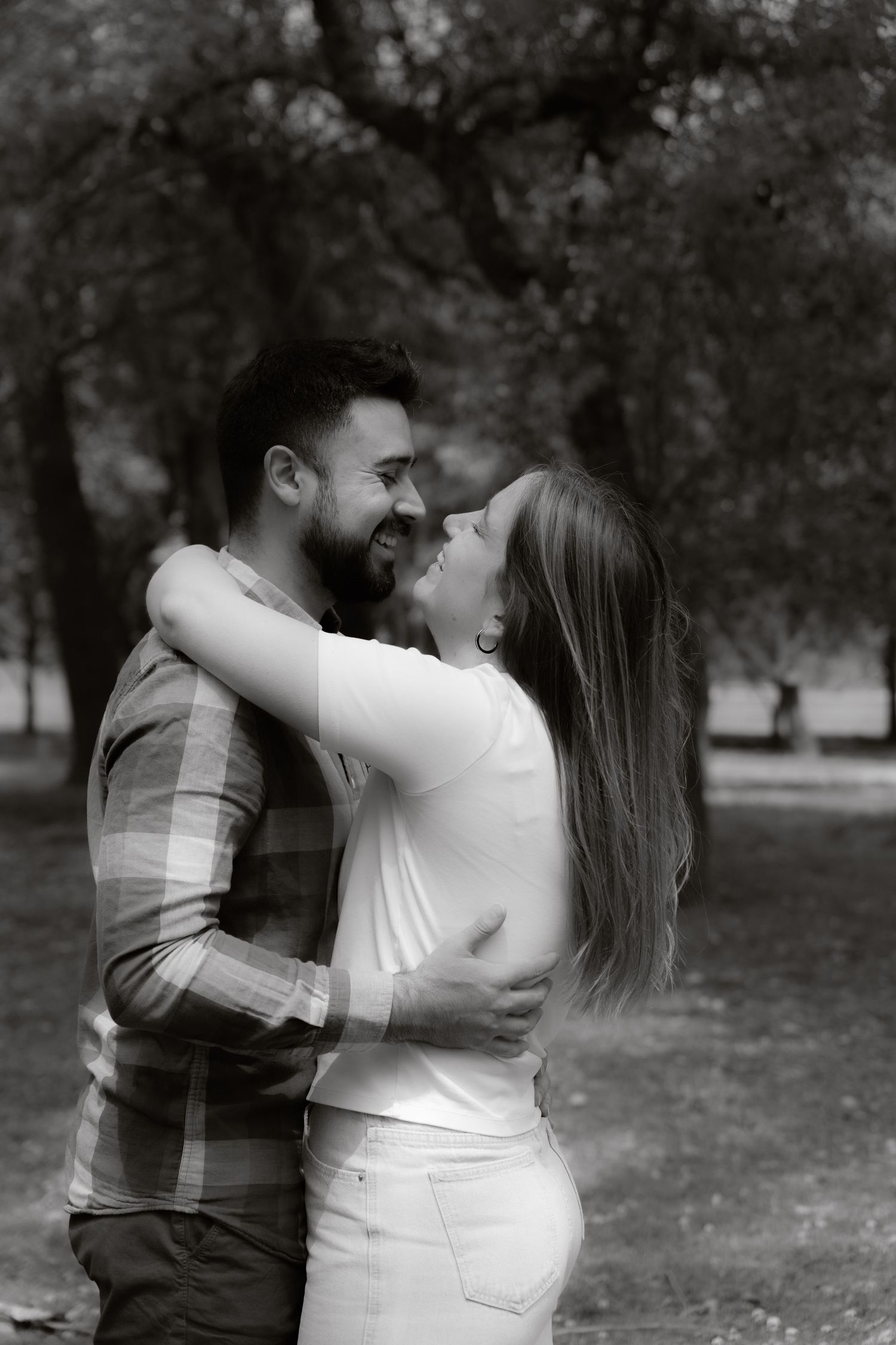 Black and white photo of a smiling couple embracing and looking at each other lovingly in an outdoor park setting.