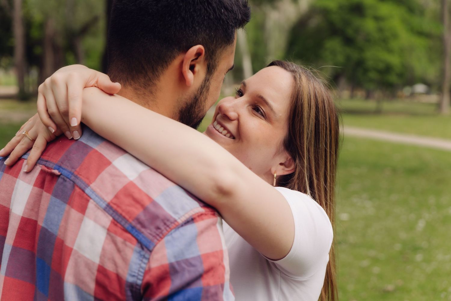 Smiling woman hugging a man in a plaid shirt from behind in a sunny green park, looking joyful.