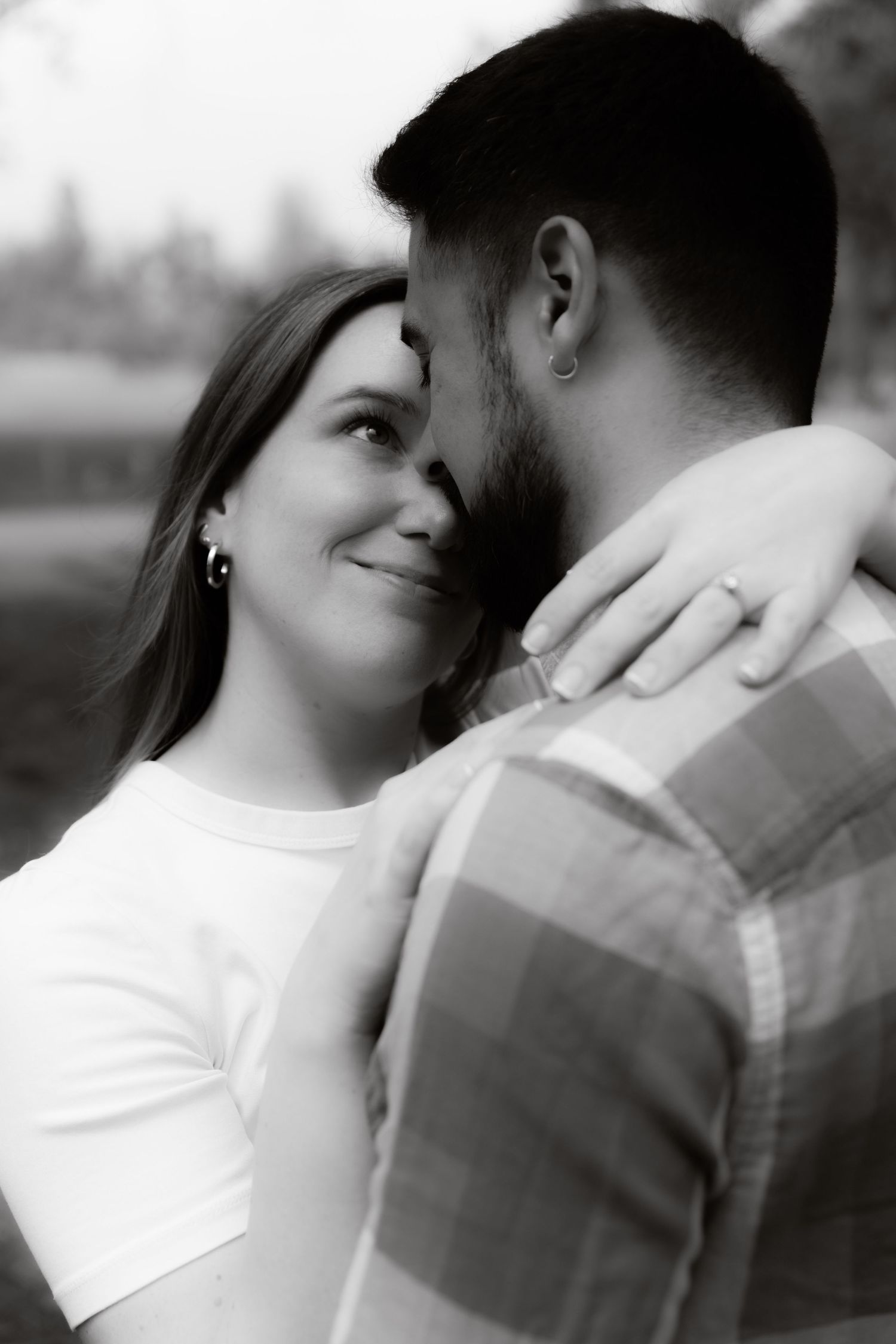 Black and white close-up of a smiling woman embracing a man, her hand resting on his shoulder in a park.