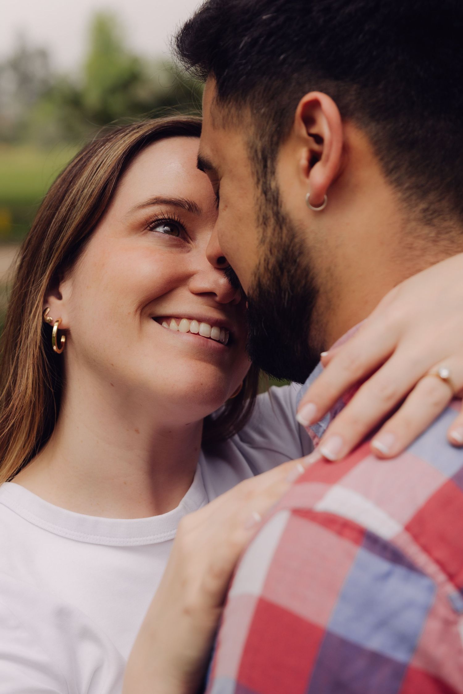 Smiling woman in white tee embraces bearded man closely, sharing a tender moment outdoors.