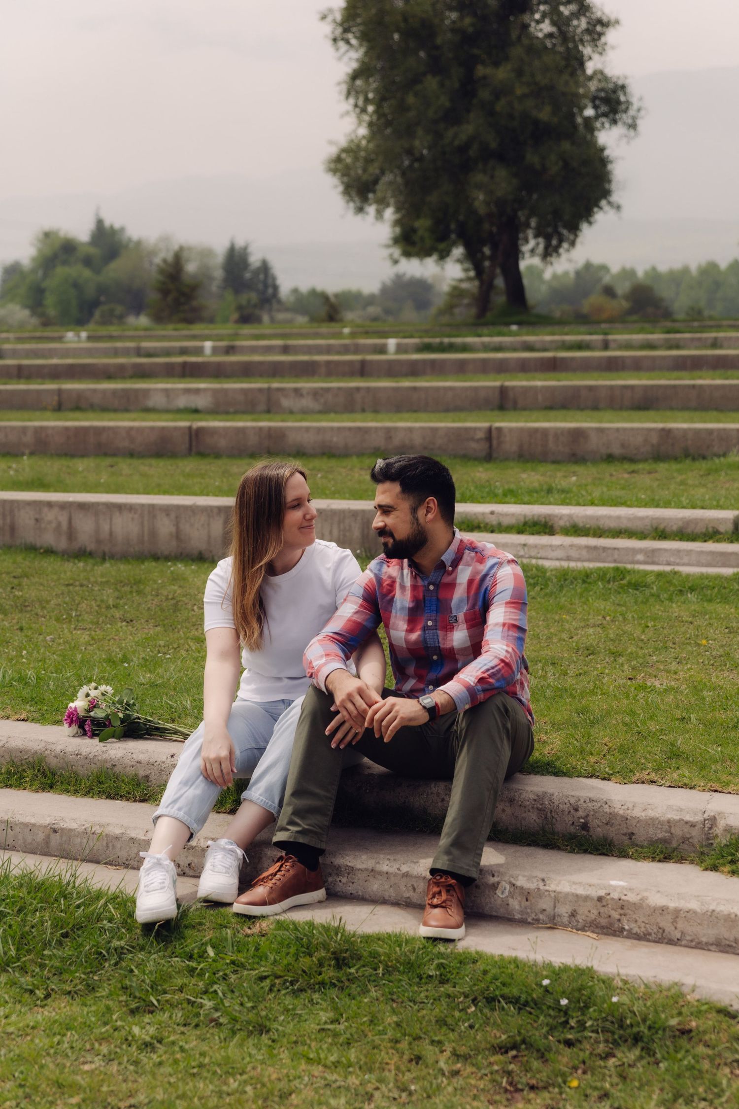 Smiling couple sitting on outdoor stone steps in a green park, gazing at each other.